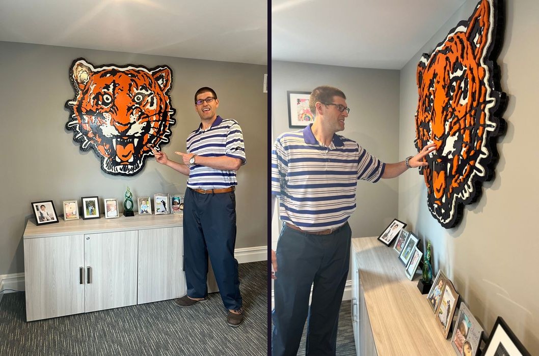 Matt Bowden pointing at the Classic Tigers tiger head cutout on his office wall, front view and a side angle