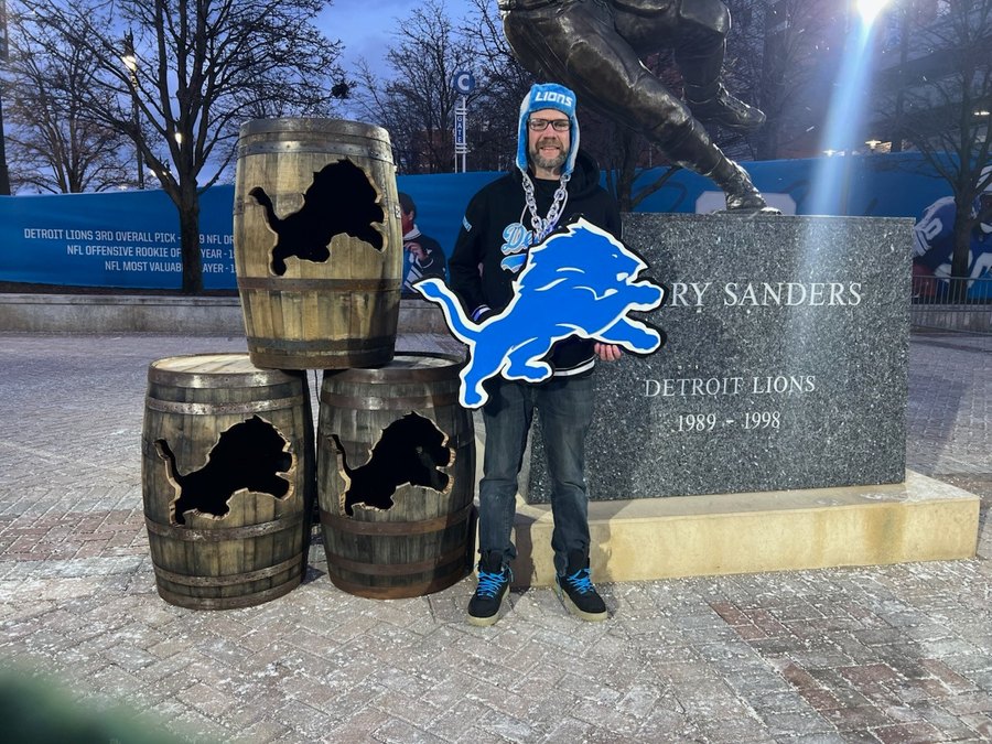 Fan with a custom Detroit Lions logo cutout outside Ford Field beside the Barry Sanders statue