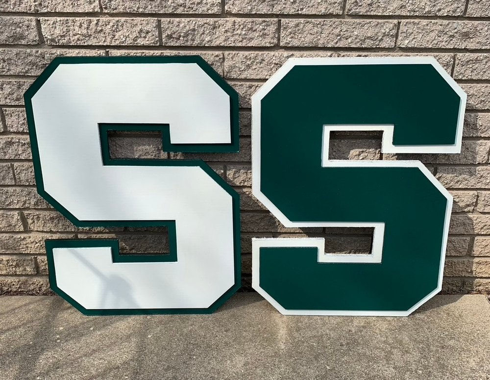 Pair of custom white and green Michigan State block S cutouts leaning on a brick wall