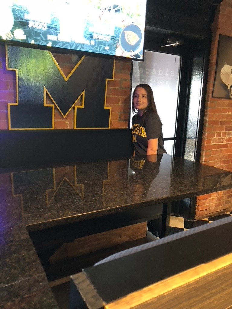 Custom navy Michigan block M cutout with maize trim on a brick wall above a granite bar top, with a patron in a Michigan shirt