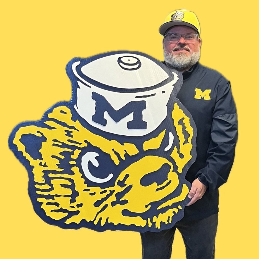 Jeremy in a maize Michigan cap holding the custom Classic Wolverine mascot cutout against a yellow backdrop