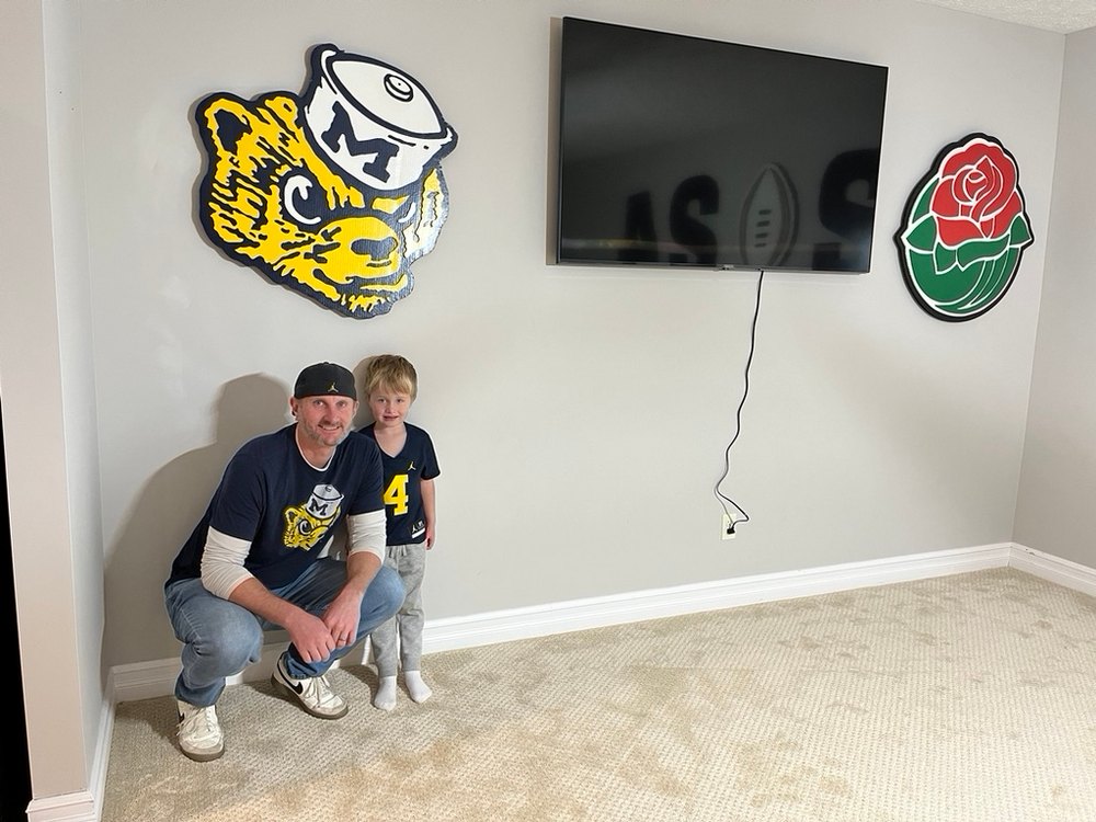 A father and young son in Michigan shirts crouching beneath the custom vintage Wolverine mascot and Rose Bowl rose cutouts on their basement wall
