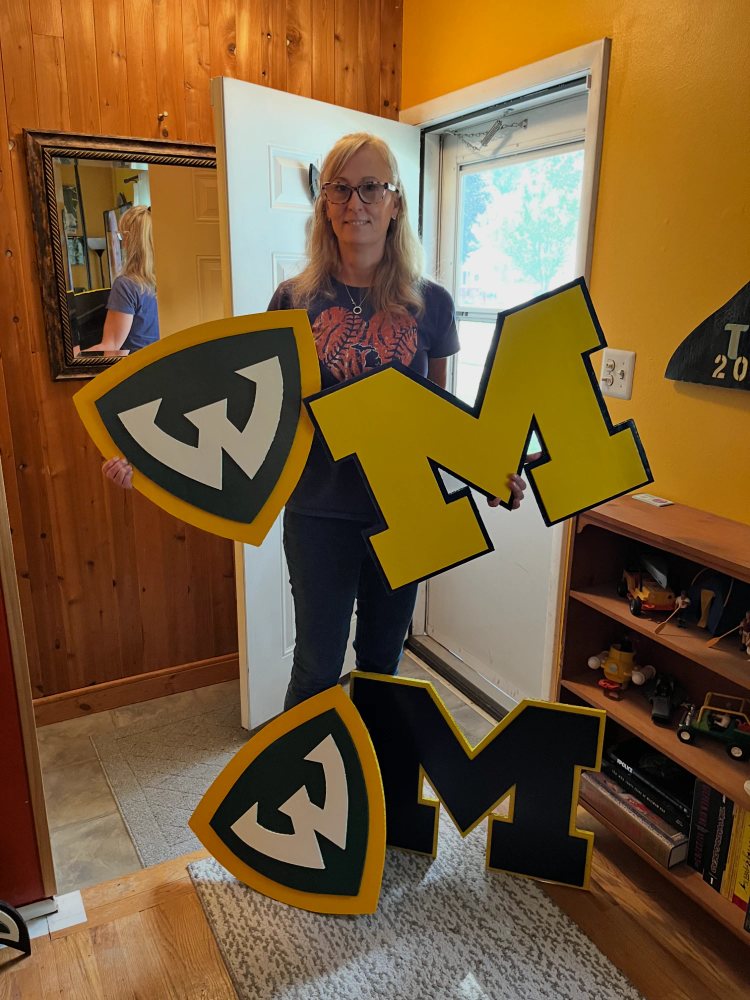 A customer holding grad gift size Wayne State University and maize Michigan block M cutouts, with a second Wayne State shield and a navy Michigan block M on the floor in front