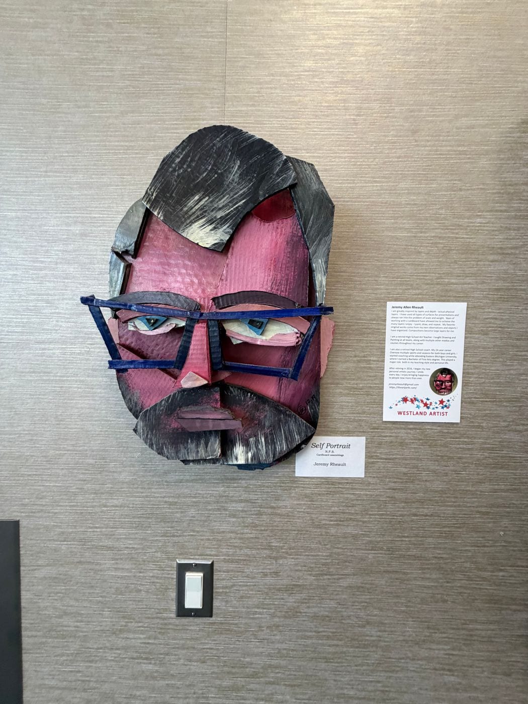 Cardboard assemblage self portrait with blue glasses, next to its label and artist statement