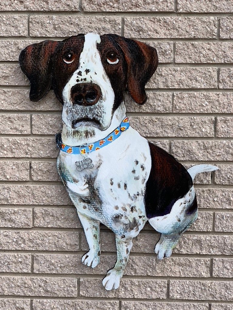 Hand-cut and hand-painted portrait of a sitting blue tick hound with a blue paw print collar and a dog tag, on a brick wall