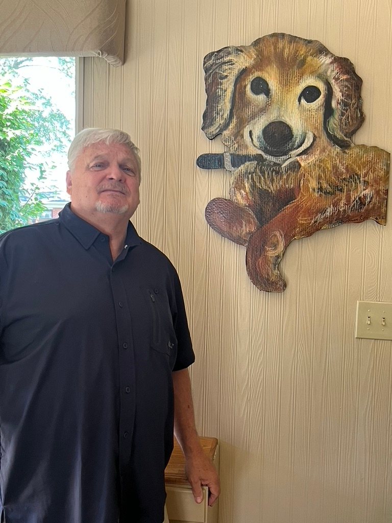 Hand-cut and hand-painted portrait of Chase, a golden dog holding a blue toy, hanging on a wood paneled wall beside the owner