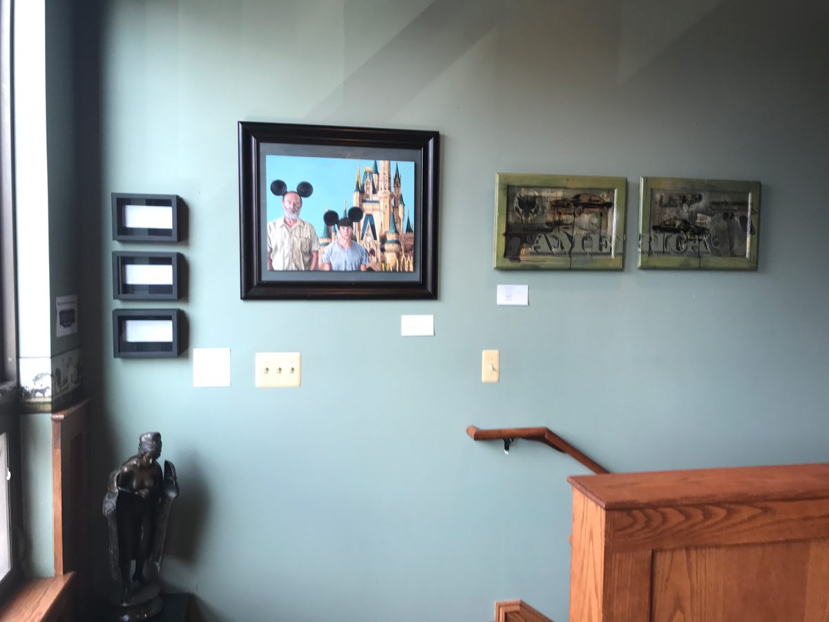 Framed photograph of two visitors with mouse ears in front of a castle, and two small framed pieces, on a sage-green wall beside the stairs