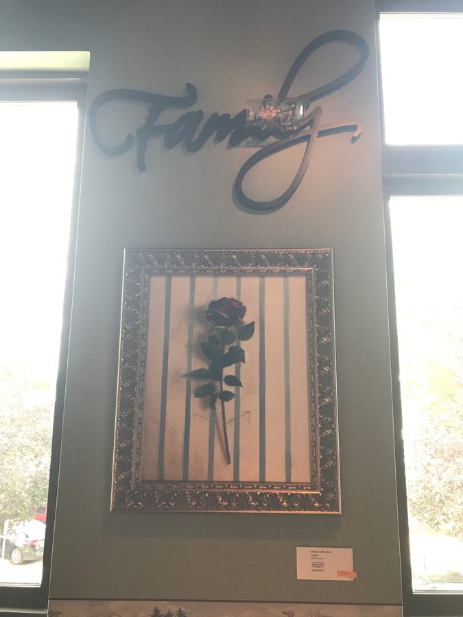 A single dark rose behind vertical bars in an ornate frame, hung under a script sign reading Family between two tall windows