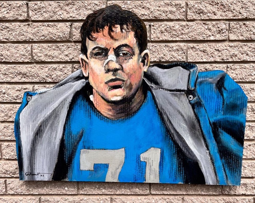 Hand-cut and hand-painted portrait of Detroit Lions Hall of Famer Alex Karras, number 71, in a blue jersey under a gray jacket, on a brick wall