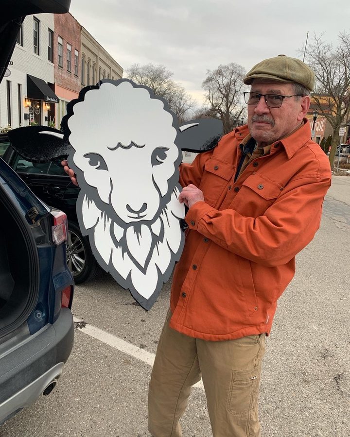 Bill Keough, in an orange jacket and flat cap, holding the white and black hand-cut Bearded Lamb sheep logo on a Plymouth street