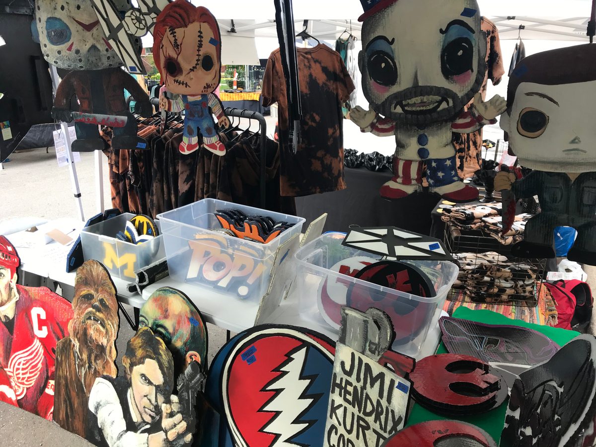 Table at the booth with hand-cut Funko-style Jason, Chucky, Captain Spaulding and Michael Myers cutouts, Chewbacca and Han Solo, a Scotty Bowman portrait, a Red Wings player, a Grateful Dead logo, a Jimi Hendrix sign and a Rebel Alliance logo