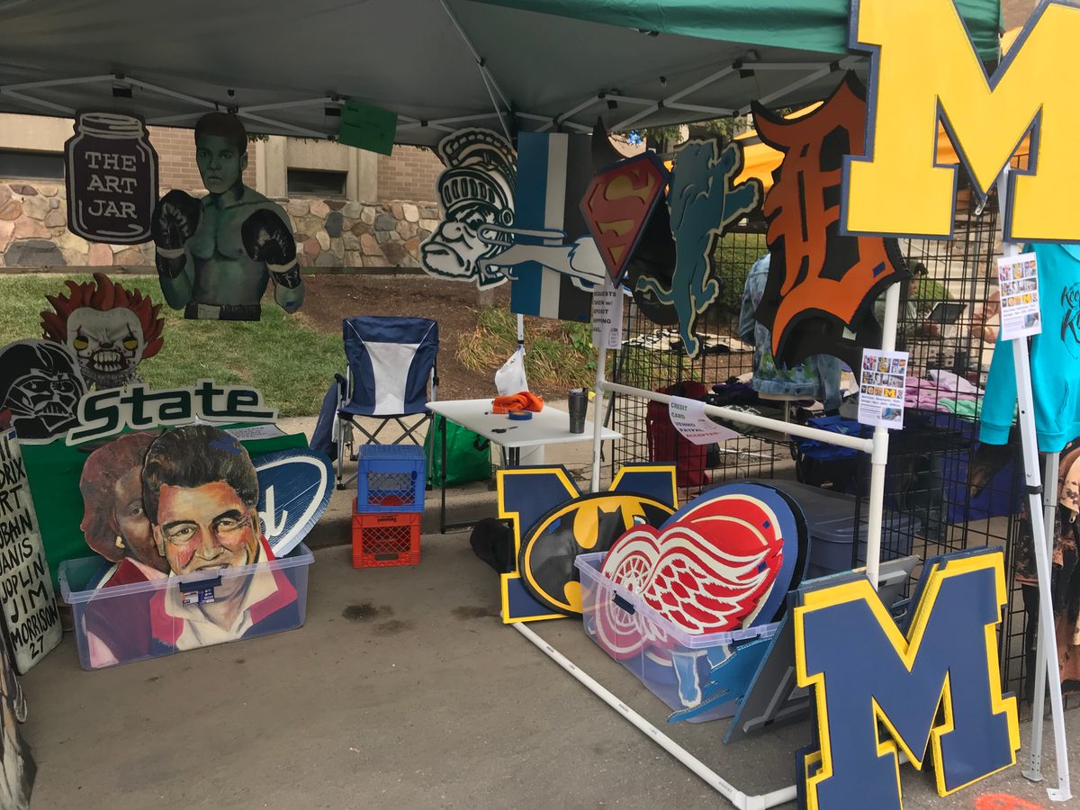 Art vendor booth under a tent with a Michigan M, Michigan State, Detroit Red Wings, Batman and Superman cutouts and The Art Jar sign