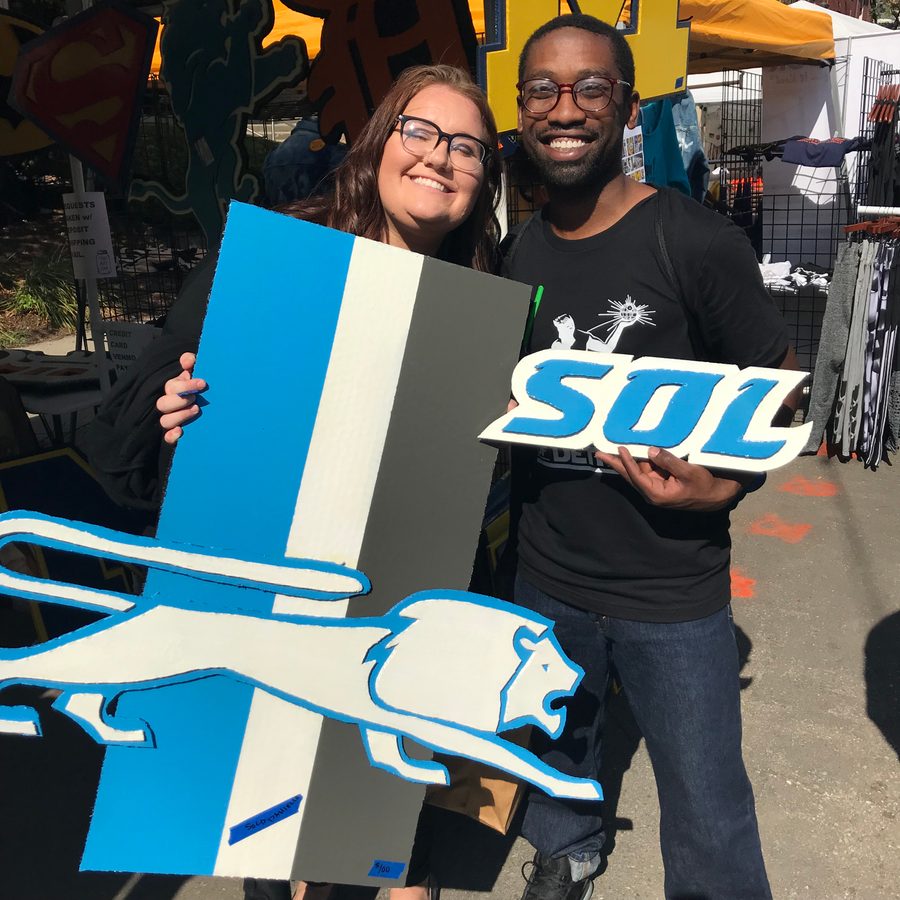 Two customers smiling and holding a Detroit Lions logo piece and an SOL sign at the booth