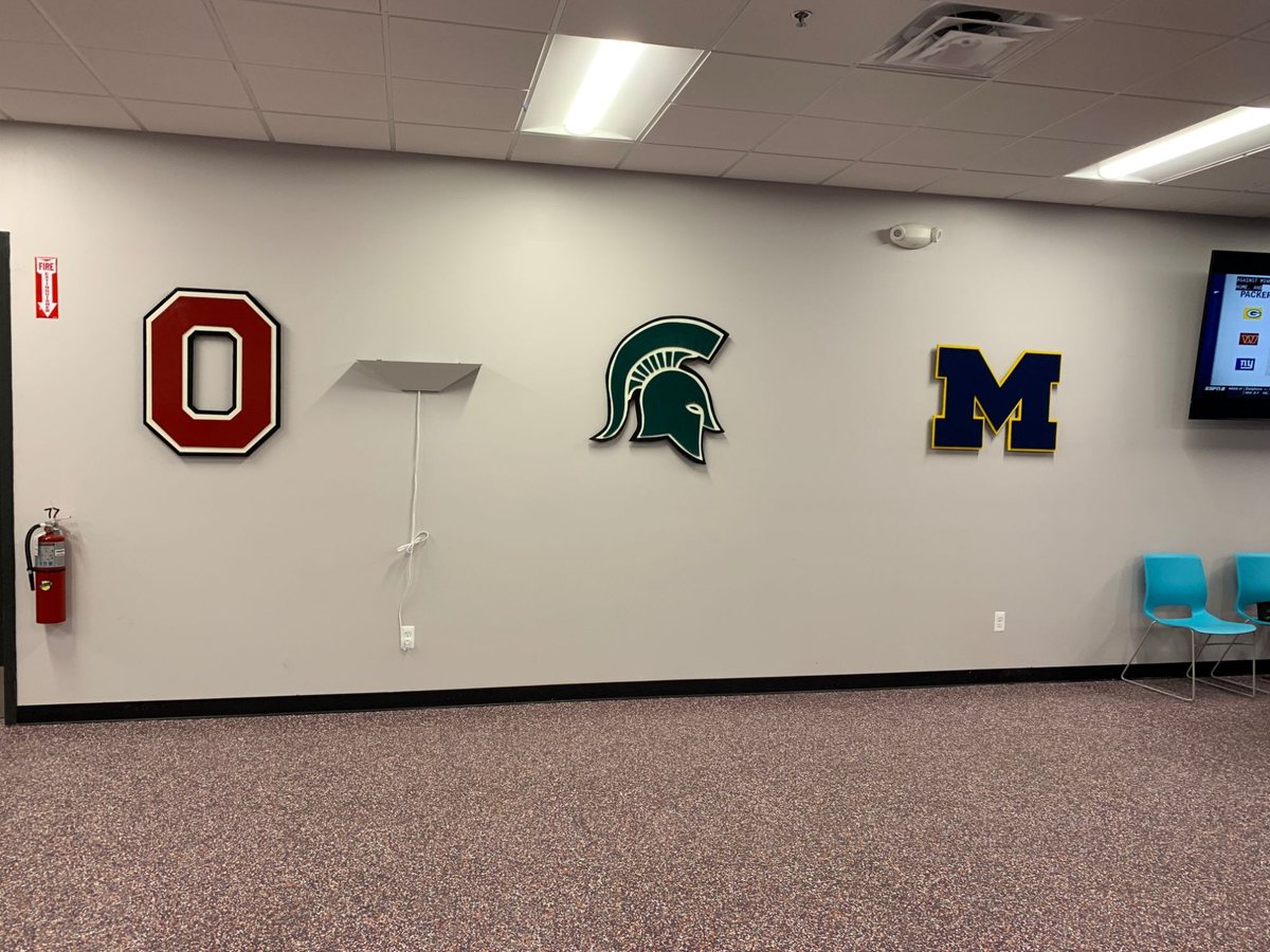 Gray wall at FedEx Ground with Ohio State O, Michigan State Spartan and Michigan M logo cutouts, a TV and a fire extinguisher