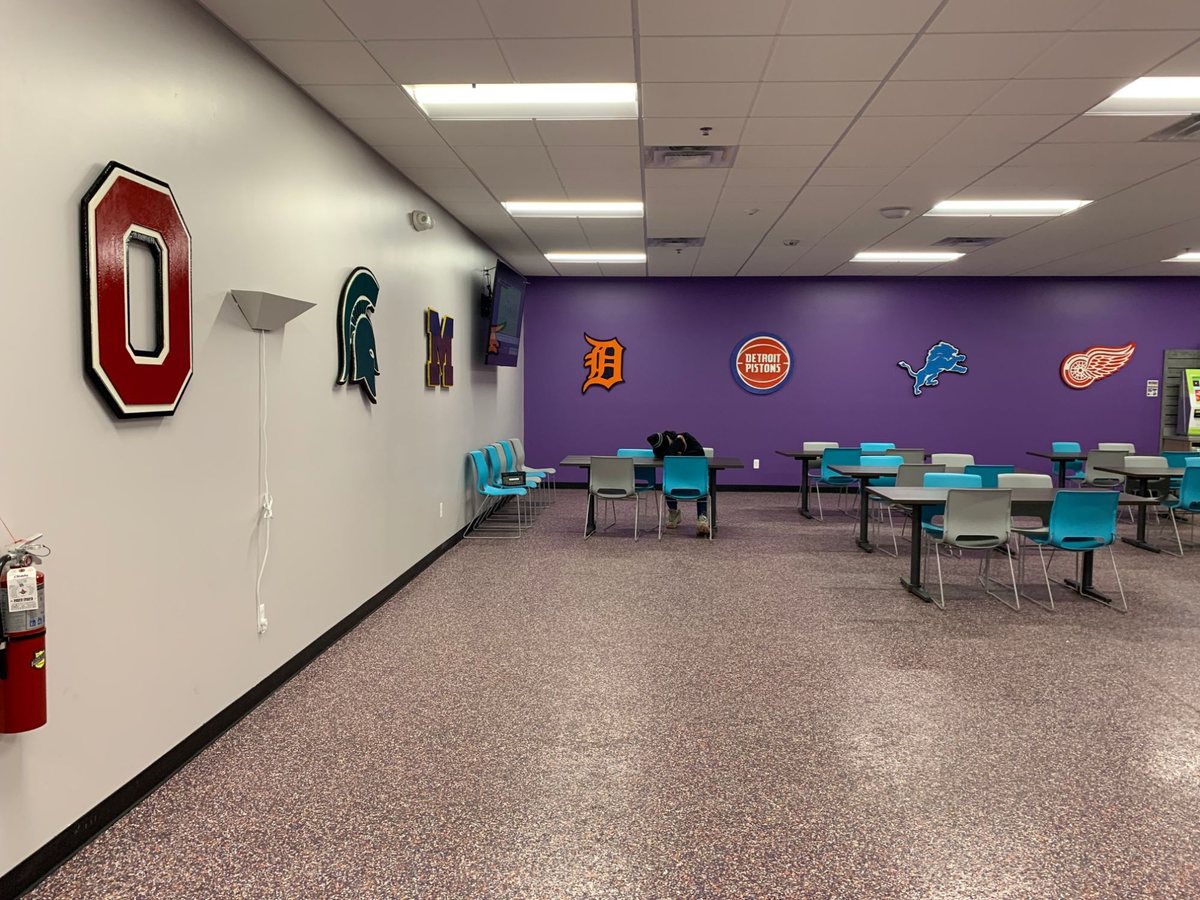 Wide view of the FedEx Ground break room with the college logo wall on the left and the purple pro logo wall at the back, tables and chairs in between
