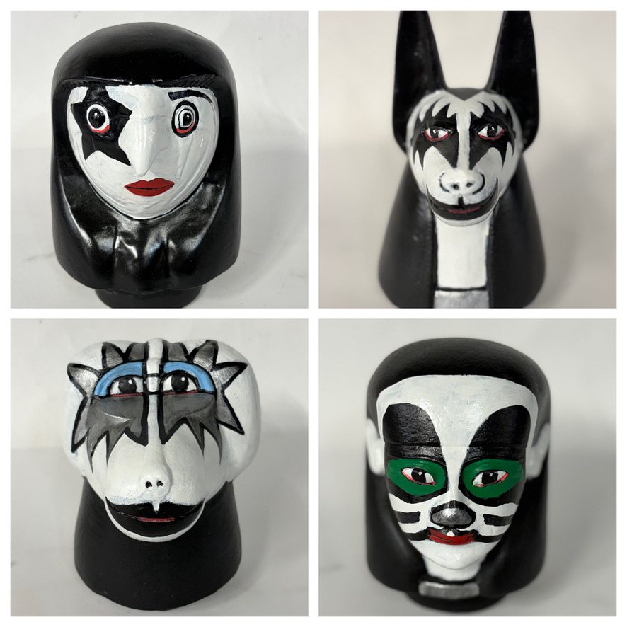 Four hand-painted KISS canopic jars, each with a black head and base: Paul Stanley with the star eye, Gene Simmons as a jackal with fangs, Ace Frehley with the silver star face, and Peter Criss with the cat face