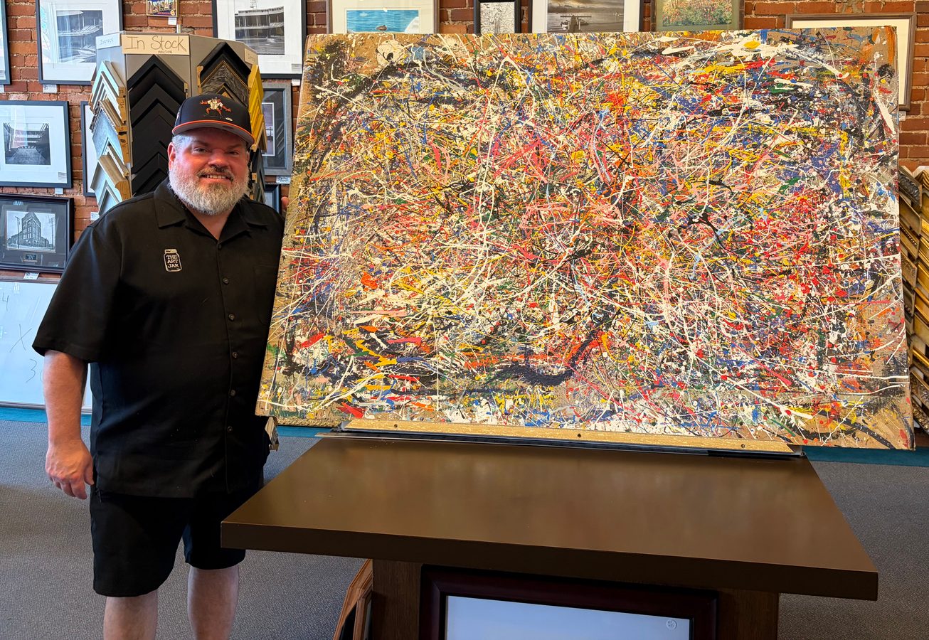 Jeremy Rheault standing beside the framed splatter painting Persevere resting on a table at Frame Works