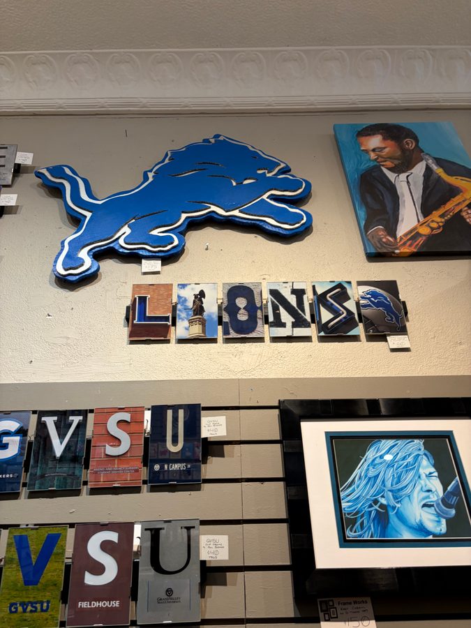 Detroit Lions logo cutout above LIONS letter tiles and GVSU, VSU letter tiles at Frame Works with a saxophone painting and a blue portrait