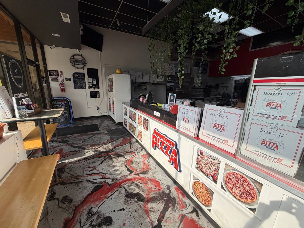 Wide view inside Hometown Pizza looking along the counter, with the hand-cut Hometown Pizza sign, pizza photos on the counter front, boxes, and a marbled red and black floor