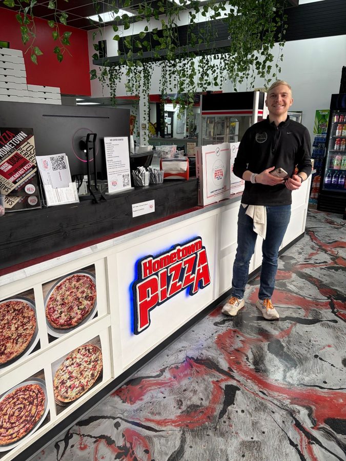 A man standing beside the counter at Hometown Pizza, next to the hand-cut, blue-backlit Hometown Pizza sign
