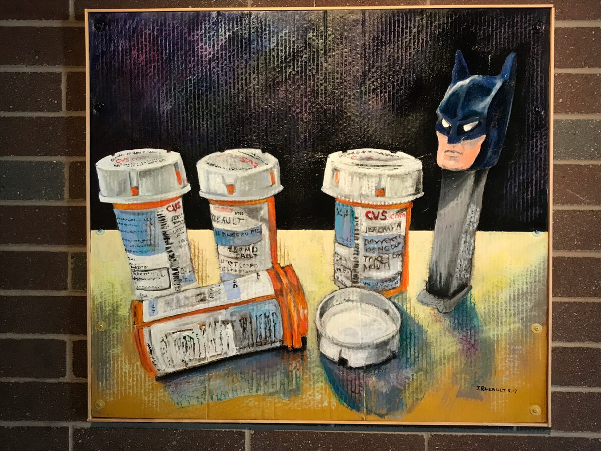 Framed painting of prescription pill bottles, an ashtray and a Batman Pez dispenser on a yellow table, hung on a brick wall