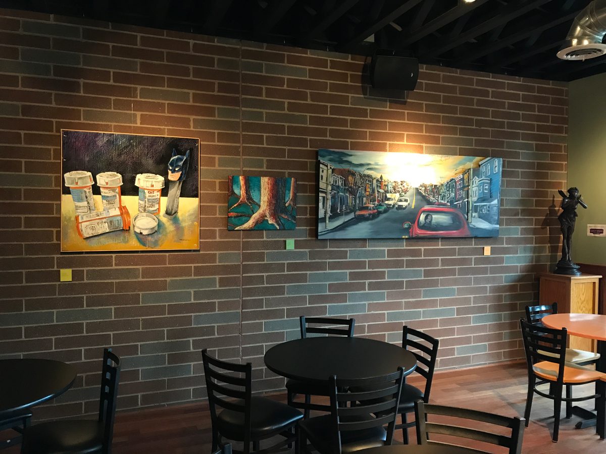 Paintings including Pills and Pez, tree roots and Street Sunset hung along the brick wall in the Up Stairs Hall
