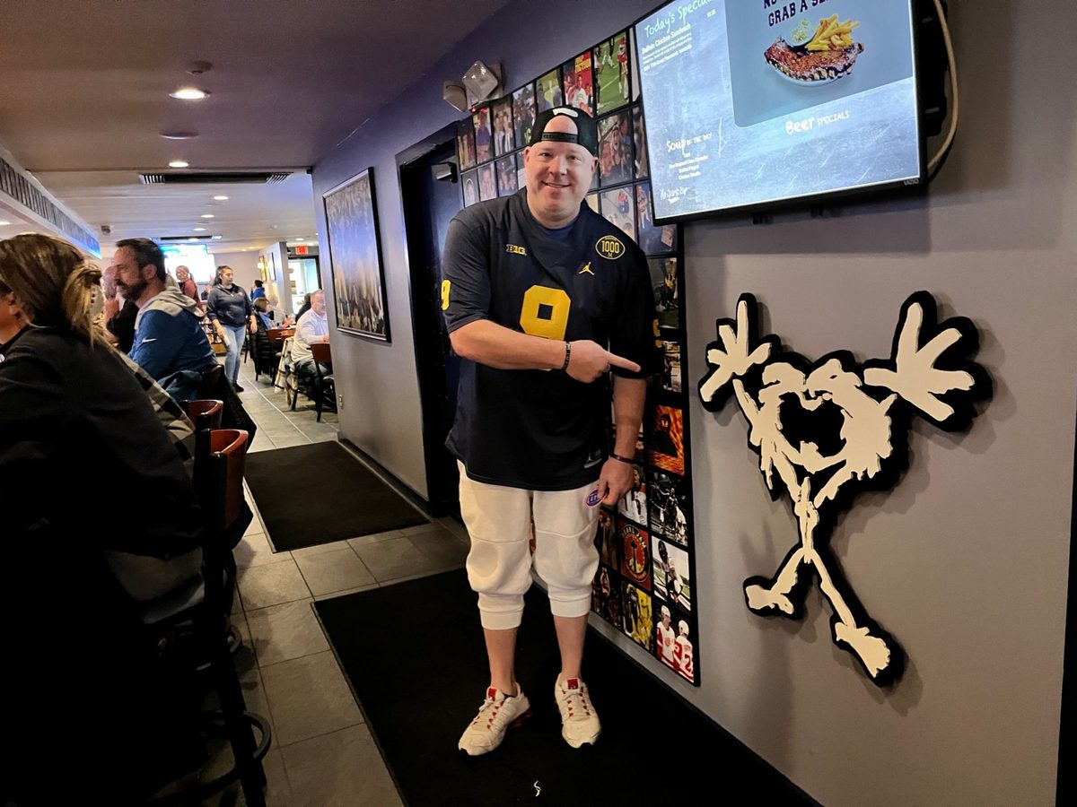 A man in a Michigan jersey pointing at the hand-cut Pearl Jam Stickman on the wall beside the photo wall