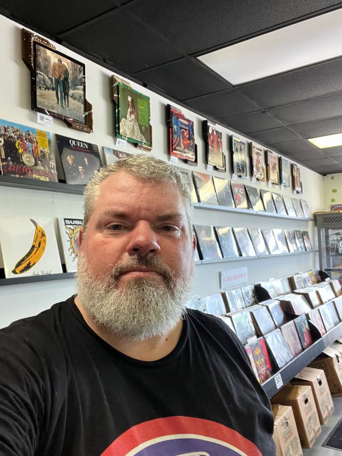 Jeremy Rheault taking a selfie in Vinyl People Records with a row of album pieces on the wall behind