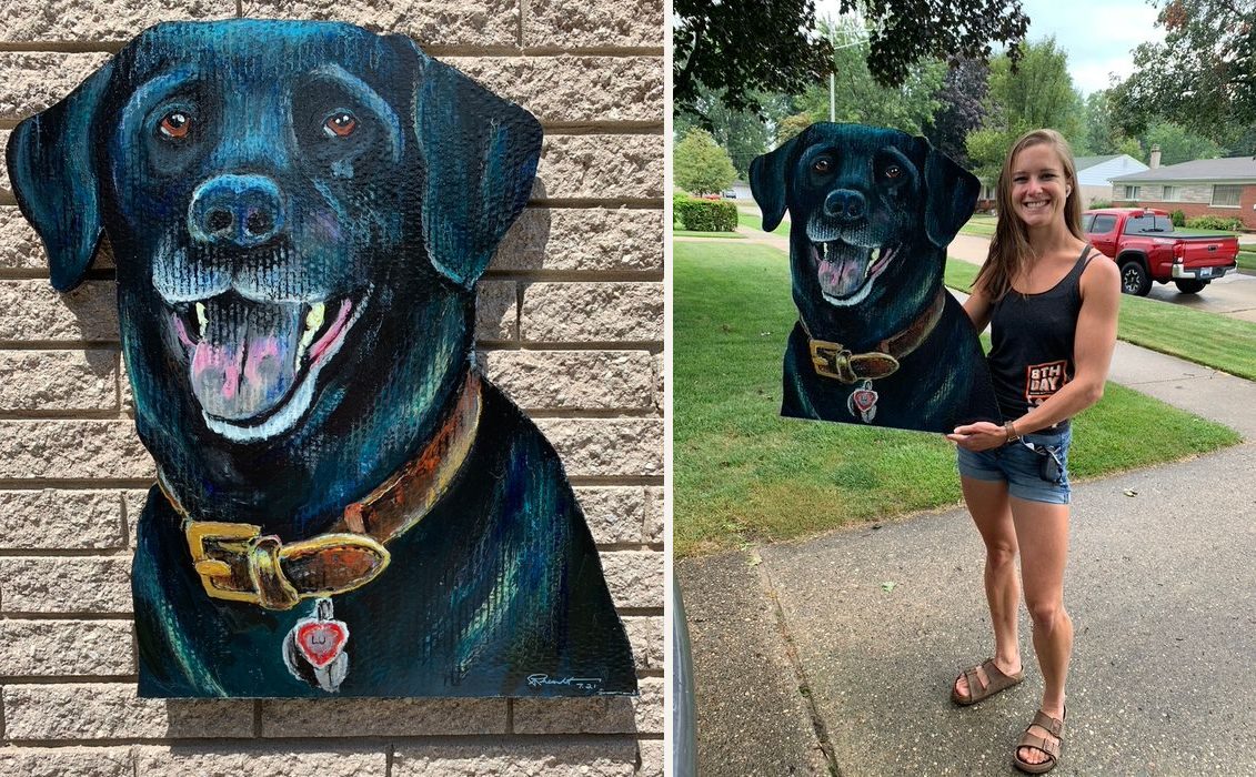 Hand-cut and hand-painted portrait of Lu, a black Labrador with a leather collar and heart tag, on a brick wall, and held by the owner in her front yard