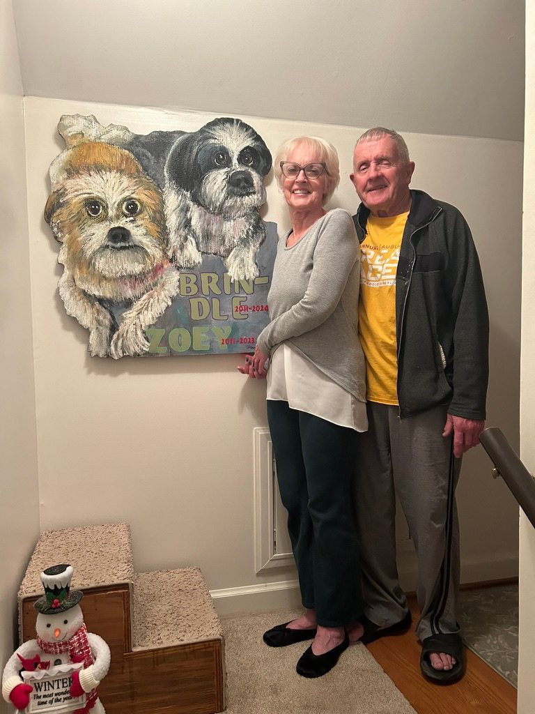 Hand-cut and hand-painted memorial portrait of two small dogs, lettered Brindle and Zoey with their years, hanging on a stairwell wall beside the owners